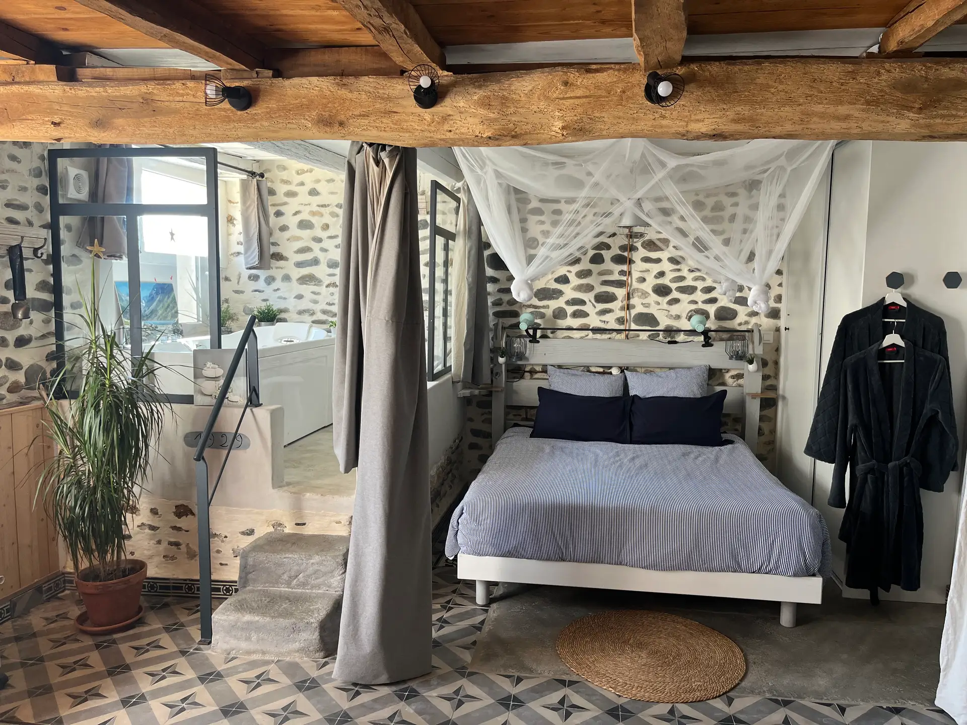 La chambre du gîte Chai Loly : lit sous moustiquaire, mur en galets, poutres apparentes, et la baignoire balnéo surélevée juste derrière.