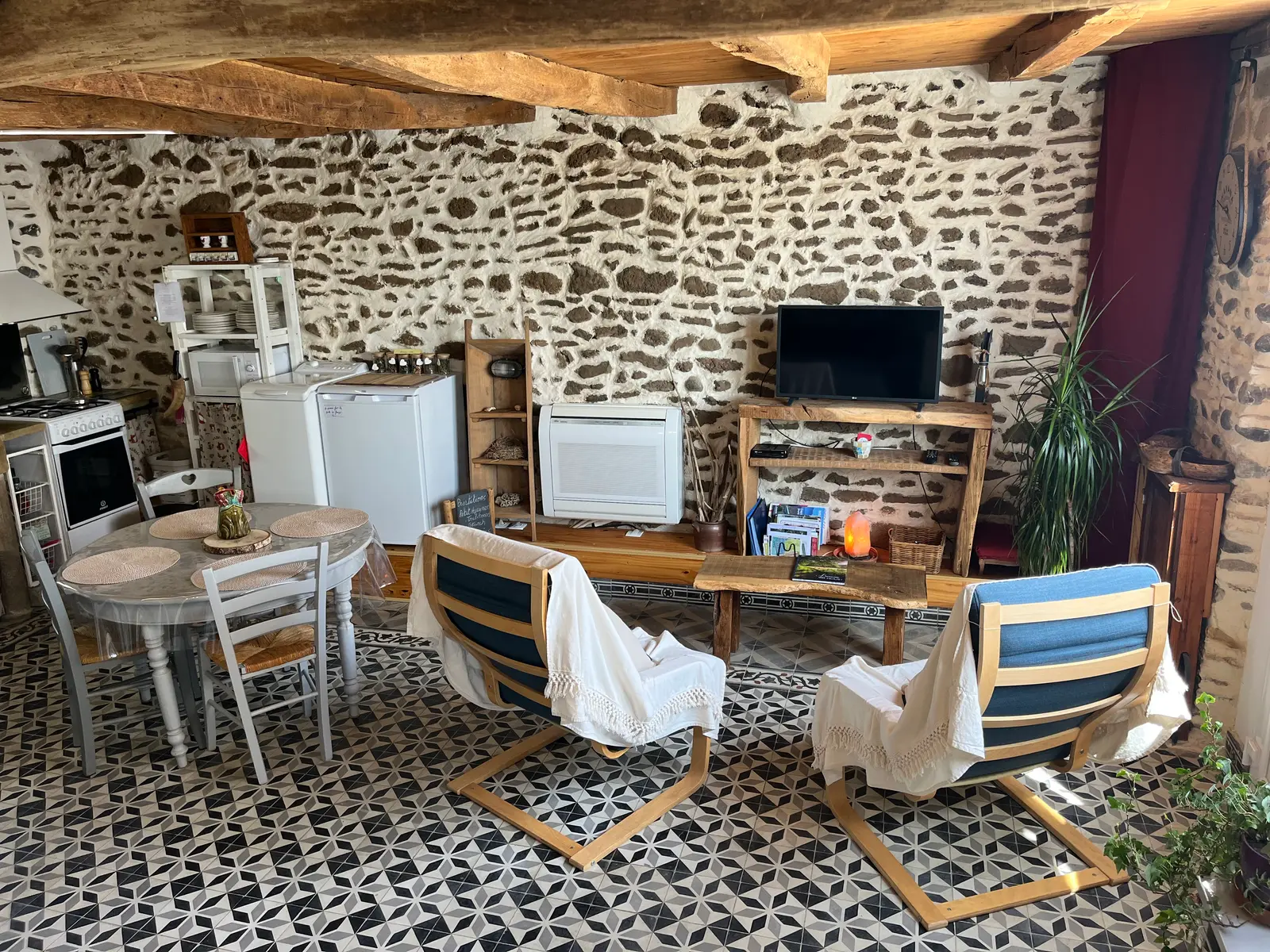 Vue d'ensemble du studio : mur en galets, poutres apparentes, carreaux de ciment, coin cuisine, table ronde et fauteuils.